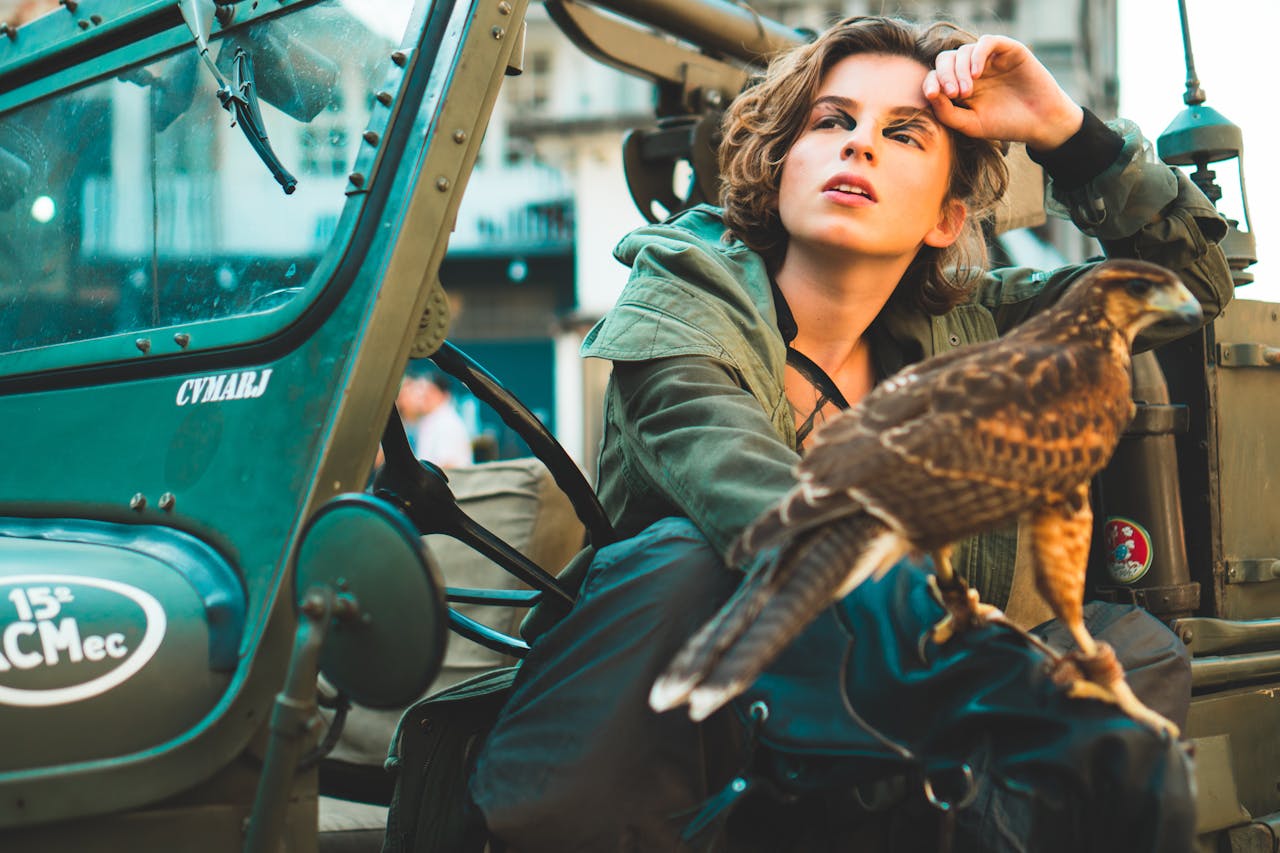 A fashionable woman posing with a falcon next to a vintage vehicle outdoors.
